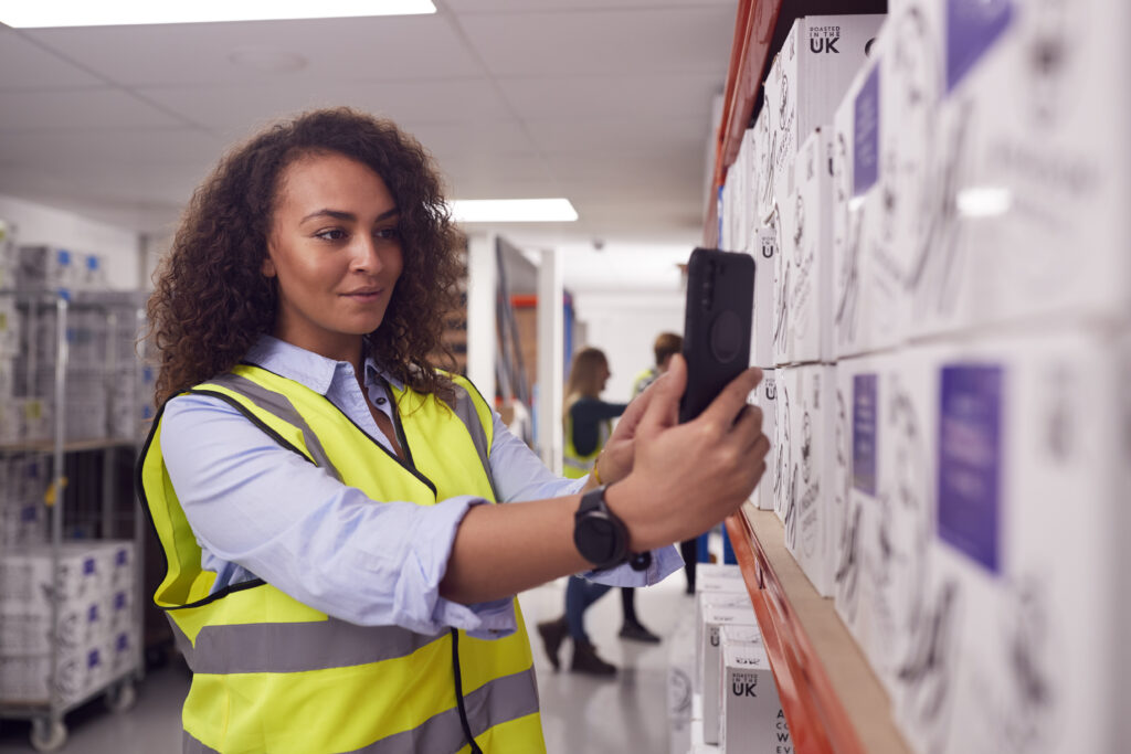 Female Worker Inside Busy Warehouse Scanning Box Barcode On Handheld Device